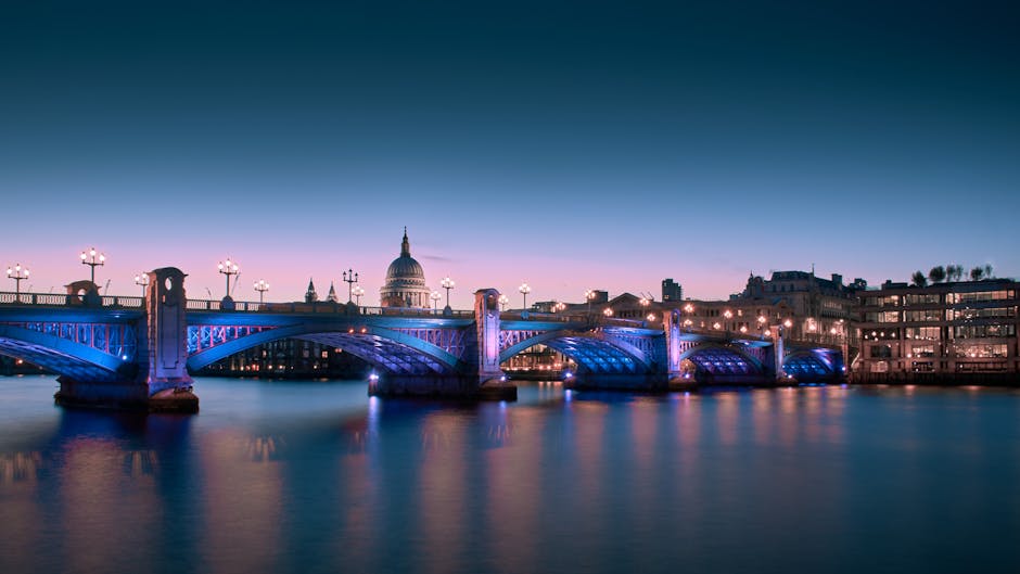 A nighttime cityscape of central London viewed from the River Thames with a well-lit bridge in the foreground, featuring street lamps and structural supports illuminated in blue and white lights. The river below reflects the lights from the bridge and surrounding buildings. In the background, the domed roof of St. Paul's Cathedral is visible with its tall spire, along with modern office buildings and historic structures. The sky above is dark but clear, emphasizing the luminous bridge and city skyline. This image captures the iconic urban scenery typical of London near Kingston upon Thames, illustrating both the vibrant night-time atmosphere and the architectural landmarks associated with the area, which is relevant for contexts involving city transport, home relocation, or professional removals in the Kingston upon Thames region.