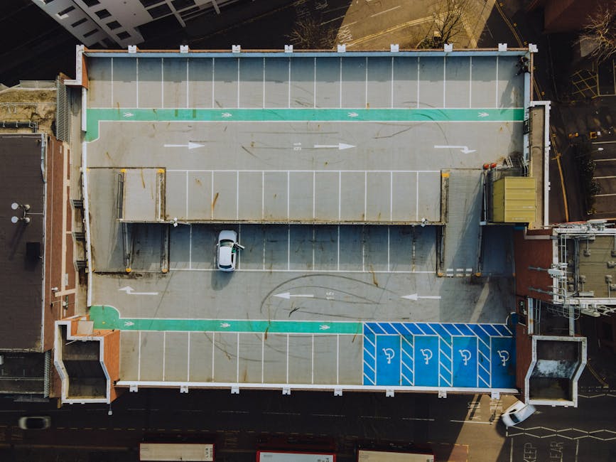 An aerial view of a flat rooftop parking area during daytime, showing designated parking spaces marked with white lines, including blue accessible parking bays near the bottom right corner. The parkin
